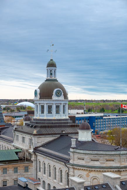 A large, historic building with a prominent clock tower featuring a dome and a weather vane on top, situated in an urban area with surrounding structures including rooftops and a modern glass building in the background. The sky is overcast with grey clouds, and a Canadian flag is visible on a flagpole near the right side of the image. In the context of house removals and relocation services, Kingston Movers might operate in such environments, involving the careful packing and transportation of household furniture and belongings. The scene highlights a combination of traditional architecture and contemporary cityscape, with open landscape beyond the city limits, relevant to the process of home relocation and furniture transport in Kingston.