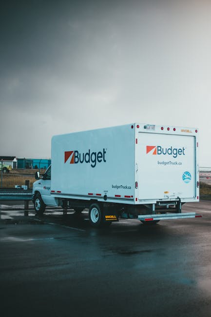 A white Budget moving van with the company logo and website address on the side is parked on a wet parking lot, likely during a home relocation or moving process. The van features red and blue branding and is positioned near an open doorway, with visible motion-blurred areas indicating recent or ongoing loading activities. Behind the van, there are industrial buildings and a partly cloudy sky, suggesting an outdoor environment typical for furniture transport or packing and moving operations. The vehicle appears to be in the process of unloading or preparing for loading, with no visible occupants in the image. This scene reflects professional removals services, such as those provided by Kingston Movers, in a setting associated with furniture transport and household relocation logistics.