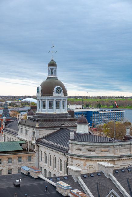 A large, historic building with a prominent clock tower featuring a dome and a weather vane on top, situated in an urban area with surrounding structures including rooftops and a modern glass building in the background. The sky is overcast with grey clouds, and a Canadian flag is visible on a flagpole near the right side of the image. In the context of house removals and relocation services, Kingston Movers might operate in such environments, involving the careful packing and transportation of household furniture and belongings. The scene highlights a combination of traditional architecture and contemporary cityscape, with open landscape beyond the city limits, relevant to the process of home relocation and furniture transport in Kingston.