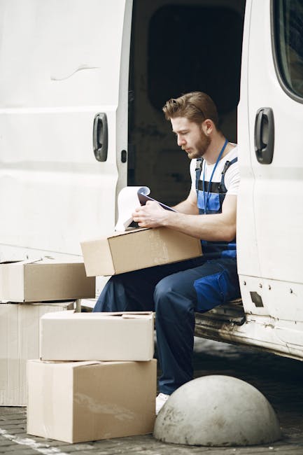 A young male removal worker with light skin and styled brown hair, wearing blue overalls and a striped t-shirt, sits on the open sliding door of a white delivery van. He is holding a clipboard and appears to be checking or recording information. Surrounding him are several cardboard boxes of various sizes, some stacked on the ground and others ready for loading or unloading. The boxes are plain and unmarked, made of brown corrugated cardboard. The scene takes place outdoors on a paved area, with a concrete bollard nearby. The van's interior is visible through the open door, showing a dark space where additional packing materials or furniture may be stored. The lighting suggests daylight, and the setting emphasizes the process of home relocation, furniture transport, or packing and moving services carried out by Kingston Movers, as seen on the webpage for same-day removals in Kingston related to the page titled 'Cheap same day removals Kingston KT1 KT2 availability and risks'.