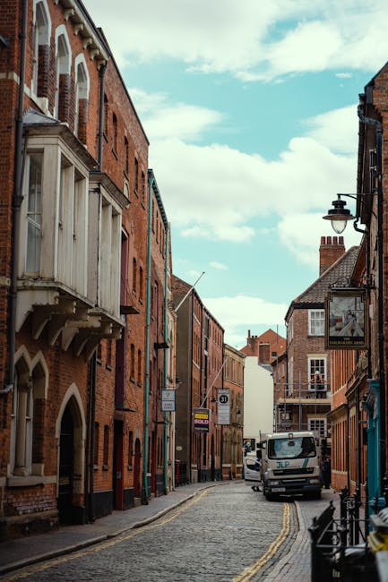 A narrow cobblestone street lined with historic red-brick buildings, some featuring decorative white timber details and bay windows. A Kingston Movers van is parked on the right side of the street, near the pavement, with cardboard boxes, wrapped furniture, and packing materials visible on the ground nearby, indicating a home relocation process. A hand truck is positioned nearby, suggesting loading or unloading activities are underway for a furniture transport or moving service. The street is illuminated by classic black street lamps mounted on the buildings, and a few pedestrians or movers may be visible nearby. The sky is partly cloudy, adding natural light to the scene, which captures the typical environment for house removals in a historic Kingston area, emphasizing the challenges of narrow access streets during furniture transport and packing and moving operations.