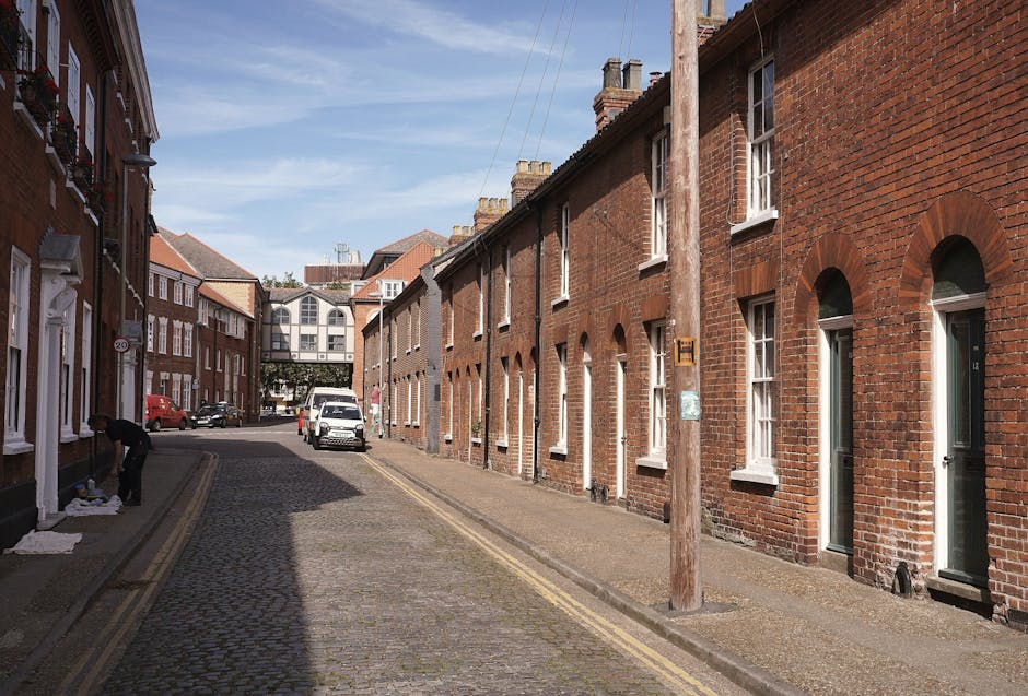 A narrow cobbled street lined with red-brick terraced houses featuring white-framed windows and doors. On the left side, a person is lifting or carrying items wrapped in white packing materials, possibly preparing for a home relocation. Several cardboard boxes and packing materials are placed on the sidewalk nearby. Parked vehicles, including a small white van and other cars, are positioned along the street. A company sign for Kingston Movers is attached to a wooden utility pole on the right side of the image. In the background, an elevated bridge or walkway connects buildings, and the sky above is partly cloudy with patches of blue. The scene depicts a typical urban residential area being prepared for furniture transport and house removals, with equipment like moving blankets and boxes present as part of the packing and loading process.