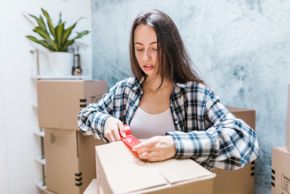 A young woman wearing a blue and white checkered shirt is seen in a room with a light grey textured wall, surrounded by multiple cardboard boxes of various sizes. She is concentrating as she unrolls red plastic tape around the top of a cardboard box, preparing it for sealing during a home relocation or packing process. In the background, there is a white shelf with a potted plant, and the flooring appears to be light-colored. The scene captures the careful packing and handling aspect of furniture transport and moving services, illustrating the importance of secure packaging to avoid mistakes on moving day, as promoted by Kingston Movers. The environment is well-lit, and the woman's focused expression reflects attention to detail in packing and loading procedures essential for stress-free house removals.