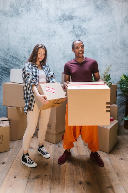 Two individuals stand inside a residential property during a home relocation process, holding cardboard boxes filled with household items. The woman on the left, dressed in a plaid shirt, beige pants, and sneakers, is carrying a box marked 'fragile' with both hands. The man on the right, wearing an orange jumper, dark trousers, and shoes, is lifting a larger box and smiling. Behind them, several cardboard moving boxes of various sizes are stacked on a wooden floor, some sealed with packing tape. A few boxes are placed near a grey textured wall, with one partially visible on the right. The environment suggests a preparation stage for furniture transport or packing and moving, with a focus on careful handling of items during loading. The lighting is bright and natural, emphasizing the organized movement of packing and logistical activities, indicative of professional removal services by Kingston Movers in a domestic setting.