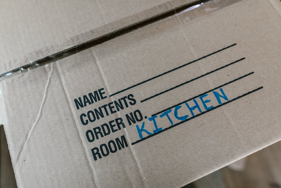 A young woman wearing a blue and white checkered shirt is seen in a room with a light grey textured wall, surrounded by multiple cardboard boxes of various sizes. She is concentrating as she unrolls red plastic tape around the top of a cardboard box, preparing it for sealing during a home relocation or packing process. In the background, there is a white shelf with a potted plant, and the flooring appears to be light-colored. The scene captures the careful packing and handling aspect of furniture transport and moving services, illustrating the importance of secure packaging to avoid mistakes on moving day, as promoted by Kingston Movers. The environment is well-lit, and the woman's focused expression reflects attention to detail in packing and loading procedures essential for stress-free house removals.