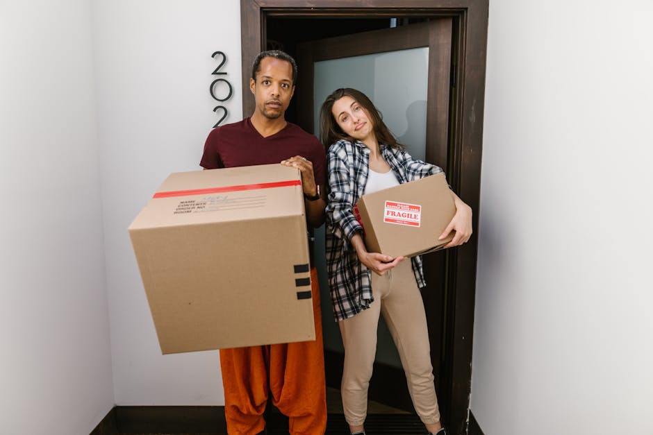 A man and a woman stand in a narrow hallway near an open doorway, each holding a cardboard box during a home relocation. The man, dressed in a maroon t-shirt and orange-brown trousers, holds a large box with black tape and packing labels, while the woman, wearing a plaid shirt over a white top and beige trousers, carries a smaller box marked with 'FRAGILE'. The background shows a wooden doorframe and a white wall with a partial view of the house number '200'. Inside the corridor, a trolley or dolly is partially visible, indicating the process of lifting and loading household items. The environment is well-lit, suggesting natural or indoor lighting, and the scene captures the logistical steps involved in furniture transport and packing during a professional removal or moving process organized by Kingston Movers, supporting efficient home removals and delivery services.