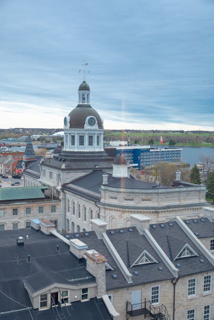 A high-angle view of a historic cityscape showing a large, ornate building with a prominent white clock tower topped by a dome and a small spire, situated on a rooftop with dark gray tiles and brick chimney stacks. Surrounding the rooftop are several smaller chimneys and dormer windows, with some rooftops covered in dark shingles. In the background, a river is visible meandering through the city, bordered by green trees and some modern buildings, including a blue construction site. The sky is partly cloudy with patches of blue, and natural daylight illuminates the scene, highlighting the architectural details of the historic building and the urban environment, as captured in a clear, detailed photograph potentially related to house or office removals, moving logistics, or building relocation services by Kingston Movers in Kingston.