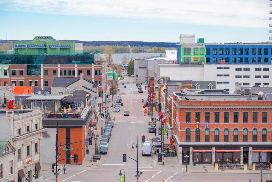 An aerial view of Kingston's High Street showing a mix of historic and modern buildings. In the foreground, a brick building features large windows and decorative architectural elements, with several colorful flags hanging from its facade. Adjacent to it, a contemporary structure with a dark exterior and a flat roof hosts outdoor seating and tables, possibly for a café or restaurant. The street is lined with parked cars on both sides, with a few pedestrians walking along the sidewalks. On the pavement, there are street lamps, traffic signals, and bollards. Further down the street, a variety of shopfronts and commercial establishments are visible, with some displaying signage and window displays. Beyond the buildings, the view extends to a body of water and green landscape in the distance, with a partly cloudy sky overhead. This setting illustrates a typical busy town centre environment, suitable for professional removals involving the packing and transportation of office or retail furniture and equipment, as undertaken by Kingston Movers.