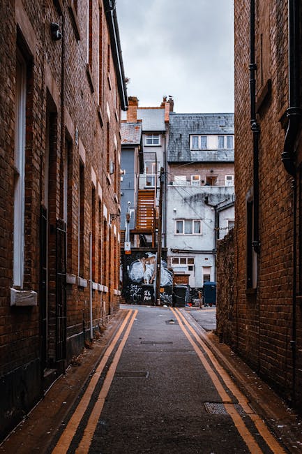 A narrow alleyway between two brick buildings in Kingston, featuring a paved surface with yellow double lines along the edges. At the far end, a vehicle is partially visible with a white plastic wrap covering items being moved. Nearby, several cardboard boxes and plastic bins are arranged on the pavement, indicating a home relocation process managed by Kingston Movers. The alley is illuminated by soft natural daylight, and external staircases and windows from surrounding buildings can be seen in the background, reflecting typical streetscape elements involved in furniture transport and packing arrangements for same-day emergency removals along Kingston’s riverside narrow streets.