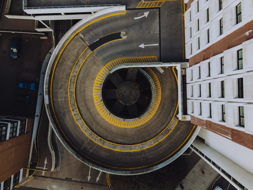 An aerial view of a circular underground parking garage with a spiral ramp painted with yellow and white striped markings, surrounded by multiple levels of residential buildings featuring white and brick exteriors. The garage's concrete surface shows signs of use, with black rubber speed bumps and directional arrows painted on the floor, guiding vehicles through the loading area. Several parked cars are visible on adjacent levels, with a narrow driveway leading to the street lined with the same yellow markings for parking and traffic flow. The lighting is natural, casting soft shadows across the scene. This image illustrates a typical urban parking structure where house removals or furniture transport might occur, with the surrounding buildings indicating a residential environment, relevant to Kingston Movers' house removal and moving logistics services.